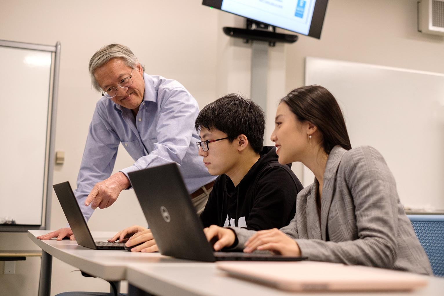 两名学生坐在电脑前的桌子前，一位教授在上海外国语大学给他们上课. 