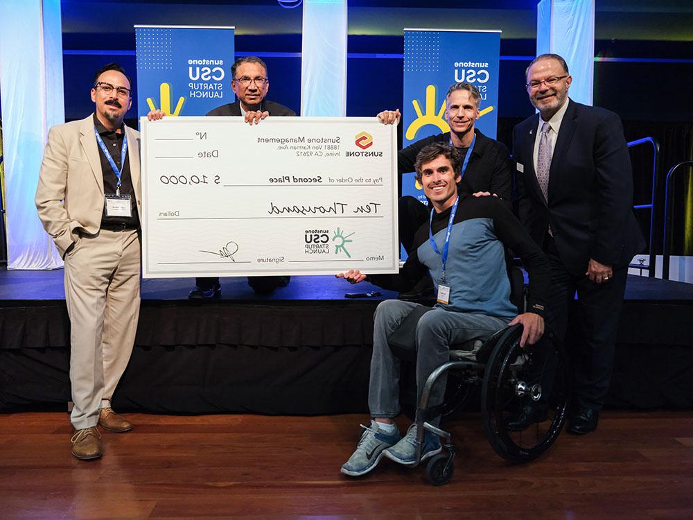 一个男, 大学年龄的创业公司创始人坐在轮椅上与两位赞助商合影, 两个主机, 还有一张25美元的大支票,000